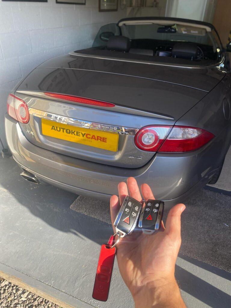 Hand holding two car key fobs near a grey convertible